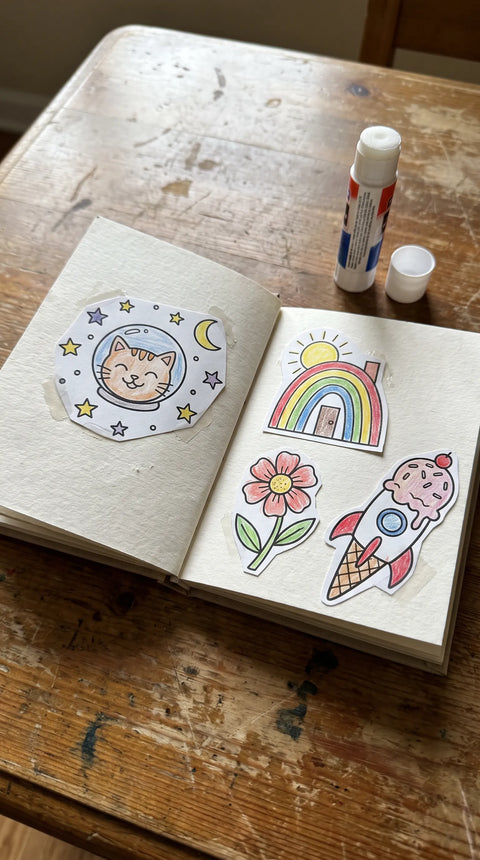 Ein kleines Heft wird mit einer Astronautenkatze, einer Blume und einem Regenbogen gestaltet.